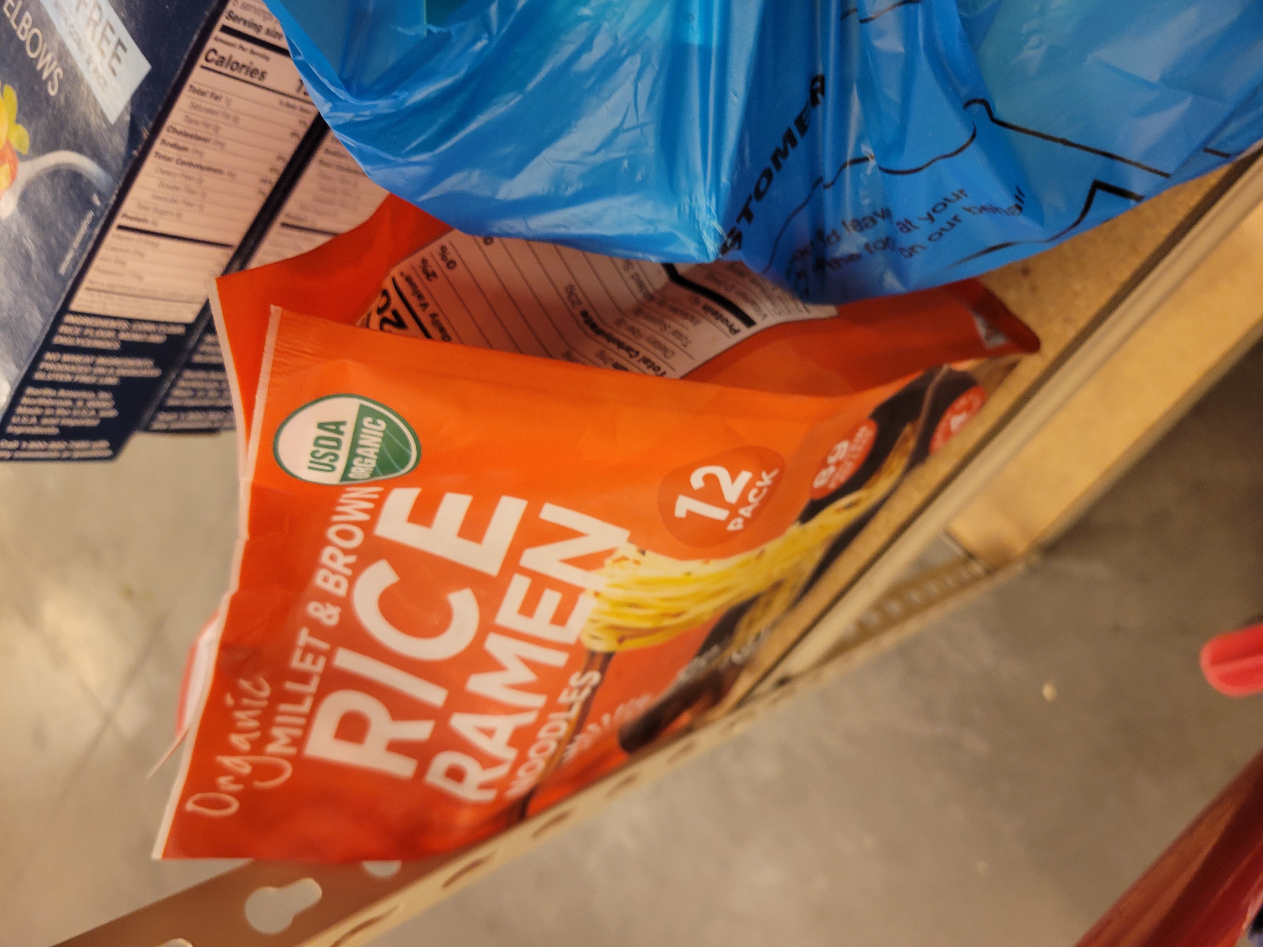 Bag of rice ramen noodles next to a blue bag and boxes of pasta behind it on a shelf.